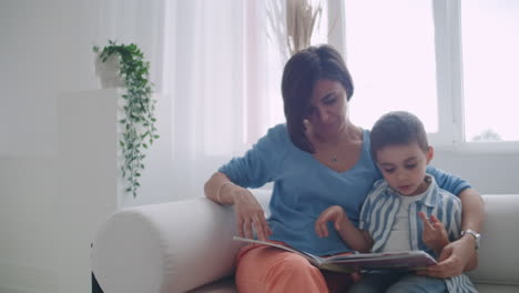 una joven madre con un niño leyendo un libro sentado en un interior blanco brillante de la casa en la sala de estar en el sofá