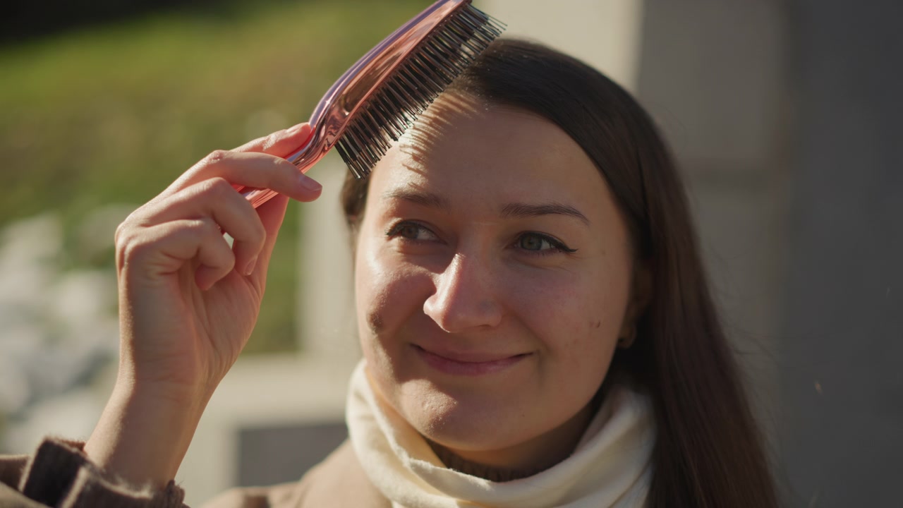 Close up of woman brushing her hair with soft lighting and natural expression. subject holds brush near long dark brown hair wearing light colored clothing in outdoor setting with greenery