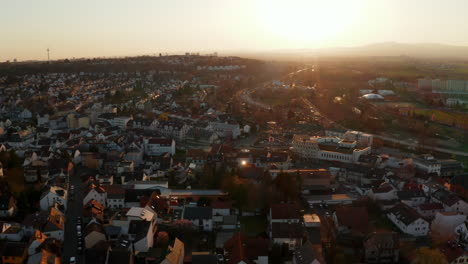 Vista-Aérea-De-La-Ciudad-Con-Ferrocarril-Y-Río-Contra-El-Sol-Poniente.-Revelando-El-Horizonte-De-La-Gran-Ciudad-En-La-Distancia.-Mal-Vilbel,-Alemania.