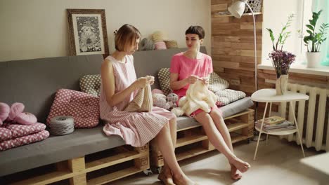 Two-girl-friends-sitting-on-couch-and-knitting-woolen-clothes