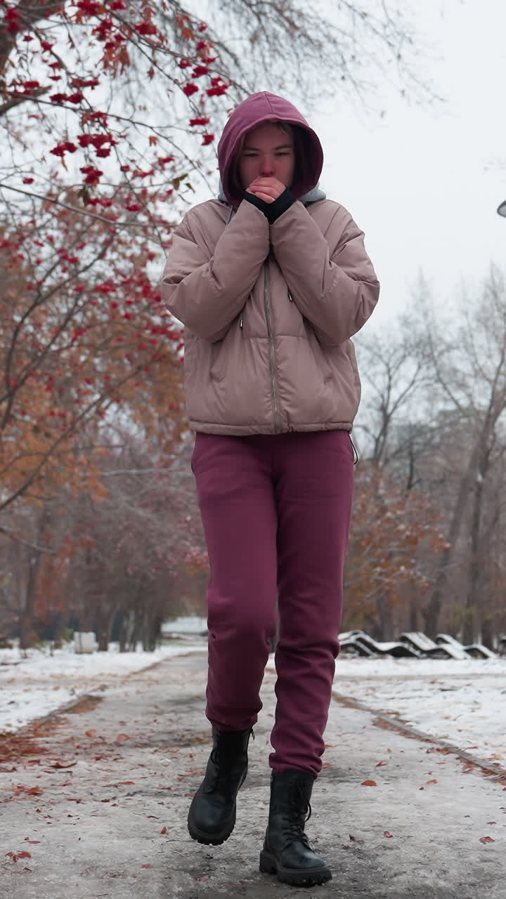 mujer con traje de invierno paseando sola, ajustando su capucha y acercando sus manos a su boca para calentarse, suelo cubierto de nieve, árboles sin hojas y hojas de otoño esparcidas