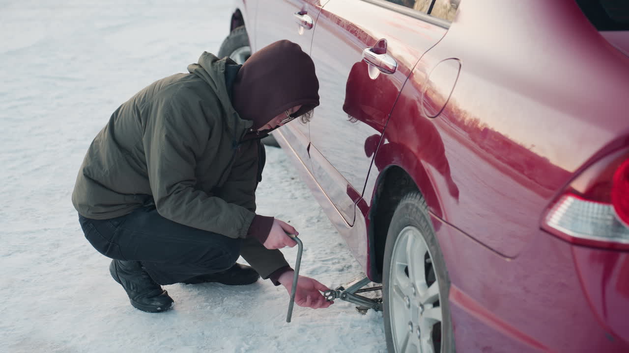 Student in winter jacket using hand to adjust jack under car with spanner in his hand on snowy ground, focused on mechanical task in cold weather