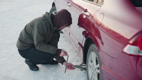 student in winter jacket using hand to adjust jack under car with spanner in his hand on snowy ground, focused on mechanical task in cold weather