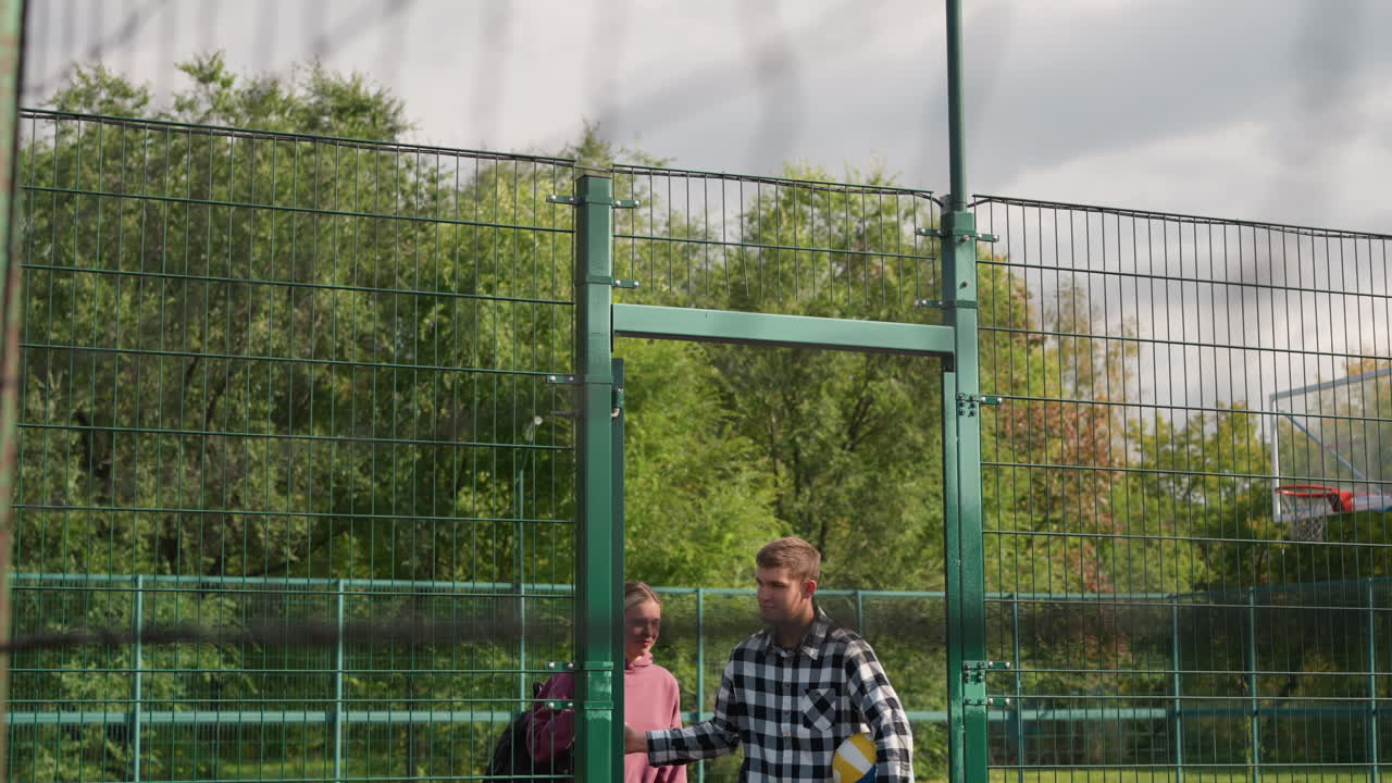 el entrenador abre la puerta verde para el entrenamiento de voleibol, el joven jugador sostiene la pelota, ambos entran en la cancha al aire libre con vegetación en el fondo, listos para el calentamiento