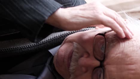an elderly gray-haired man sitting on a chair in a room by the window, his head hurts, rubbing his temples, migraine and depression. vertical video