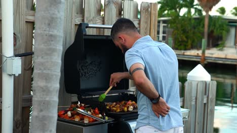 caucasian man basting chicken, beef, vegetable shish kabob skewers on waterfront charcoal grill