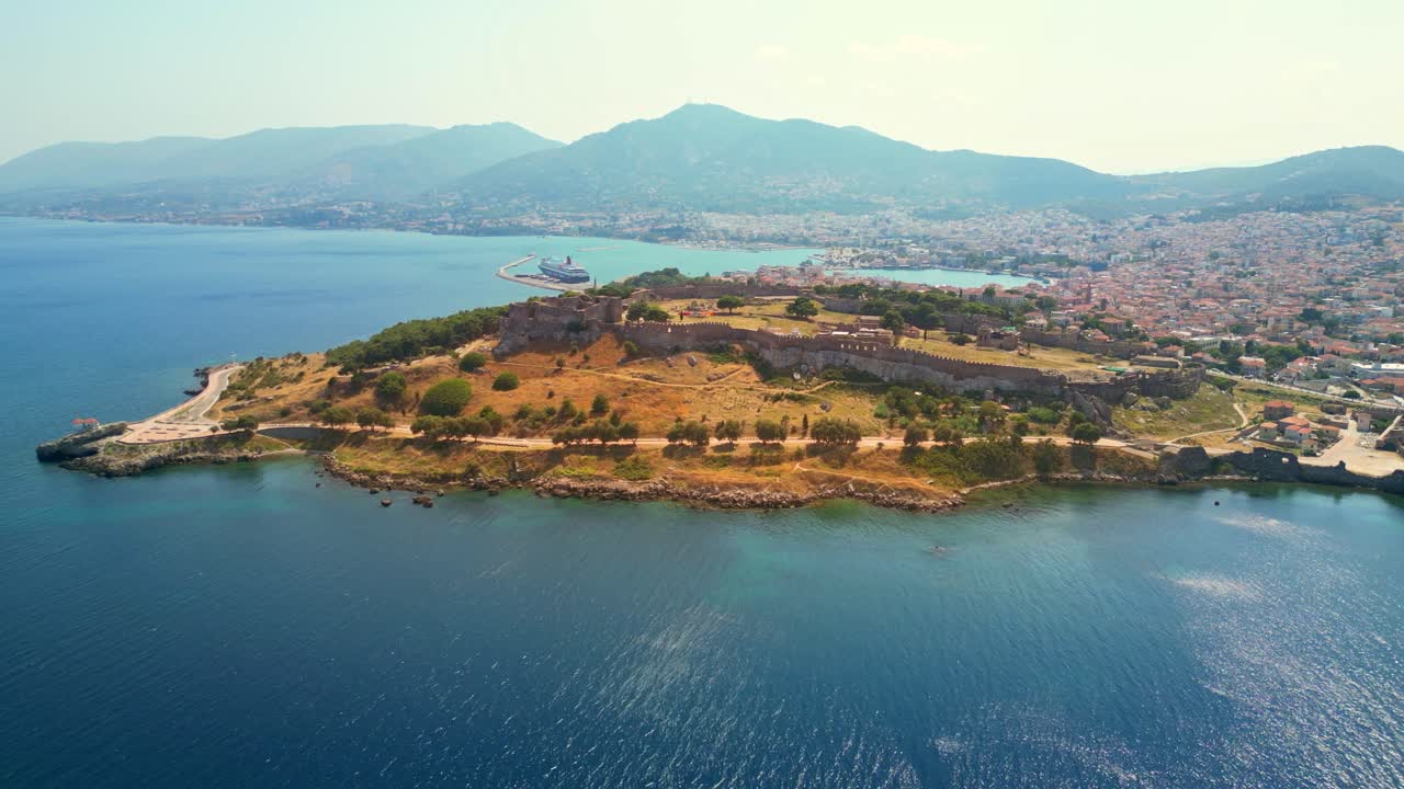 Drone shot orbiting the whole peninsula of the Castle of Mytilene, Greece, on a bright sunny day, with a view of the port, terracotta-roofed houses, and hills.
