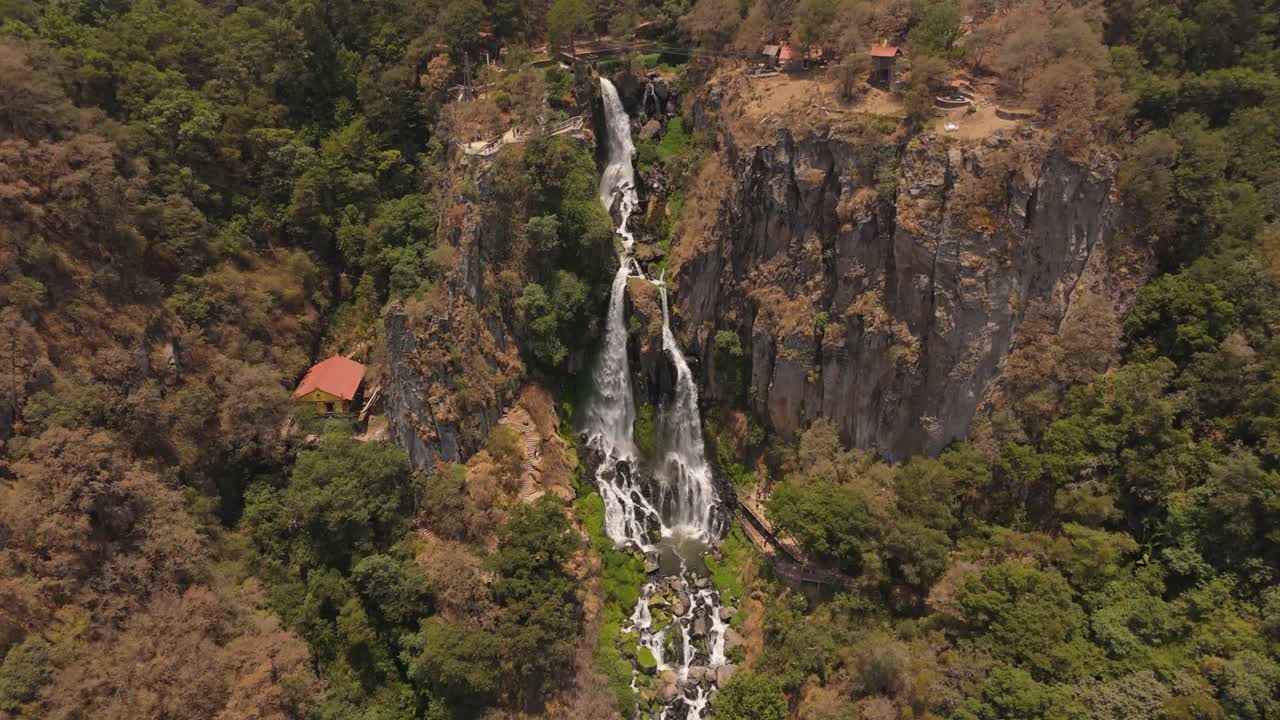 A powerful waterfall flows down a forested cliff, with quaint houses nestled in the landscape.