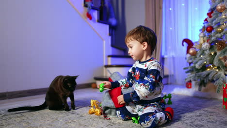 small caucasian kid sits on the floor near christmas tree. toddler pulls the candies from stocking and puts them in front of the black cat.