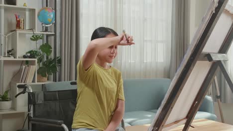 artista asiática en silla de ruedas sosteniendo un pincel limpiar el sudor antes de cruzar los brazos y sonriendo después de terminar de pintar en el lienzo