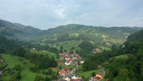 Seitlicher-Flug-über-Ein-Dorf-In-Einem-Tal-Mit-Nebel-Und-Sonnigen-Gebieten-Mit-Wiesen-Und-Vielen-Eichen-Mit-Ihren-Häusern-Und-Straßen-In-Einer-Bergumgebung-An-Einem-Bewölkten-Himmelstag-Im-Sommer-Kantabrien-Spanien