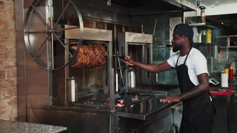 A-man-in-a-black-apron-is-spinning-a-kebab-skewer-at-a-doner-market.-A-Black-person-in-a-white-T-shirt-on-the-background-of-a-doner-market