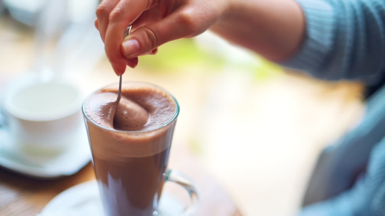 Donna con maglione blu che mescola cioccolata calda in una tazza trasparente in un bar