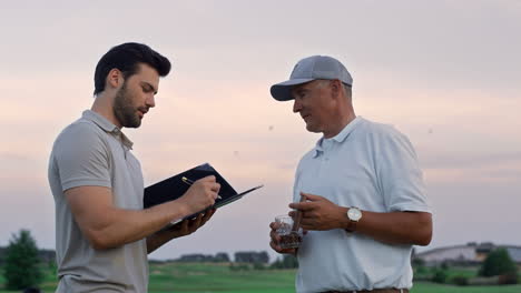 golf team chatting friendly on sunset golfing field. two men shake hands outside