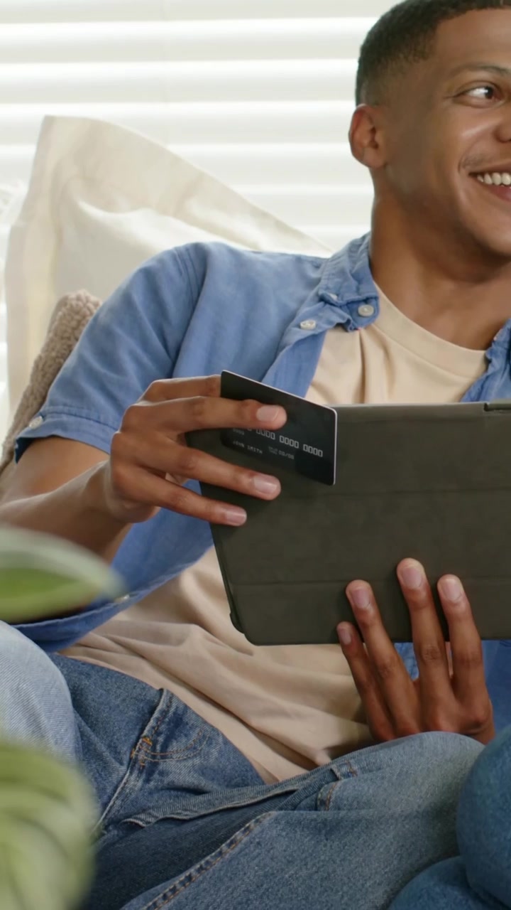 Vertical video: Camera panning showing man tapping tablet and holding card at home checking info
