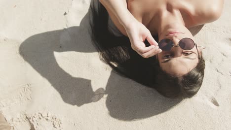 Overhead-Porträt-Einer-Glücklichen-Hispanischen-Frau-Mit-Sonnenbrille,-Die-Am-Strand-In-Der-Sonne-Liegt,-Zeitlupe
