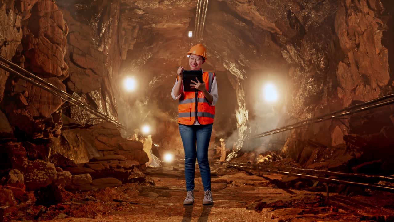 Figura intera di un'ingegnera asiatica con casco di sicurezza che prende appunti sul tablet e si guarda intorno in una galleria di miniera sotterranea