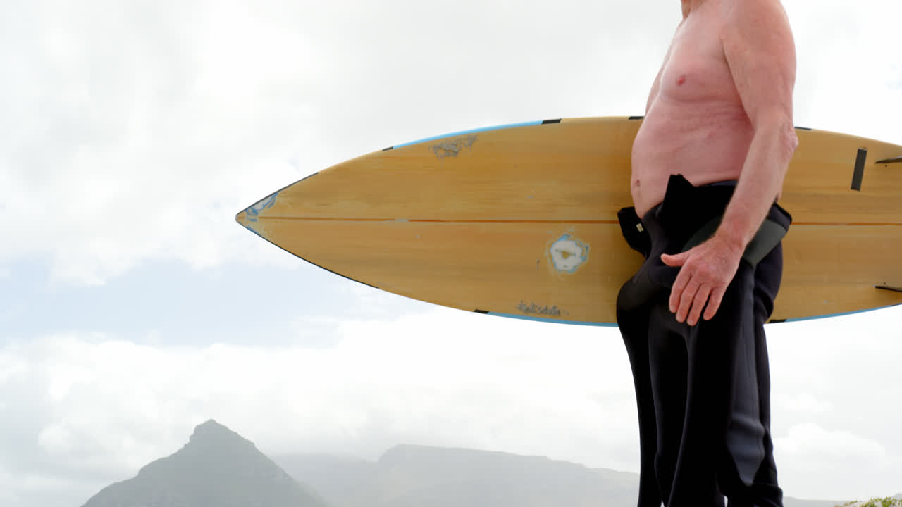 해변 4k에서 서핑 보드와 함께 서 있는  ⁇ 은 백인 남자의 낮은 각도 측면 뷰