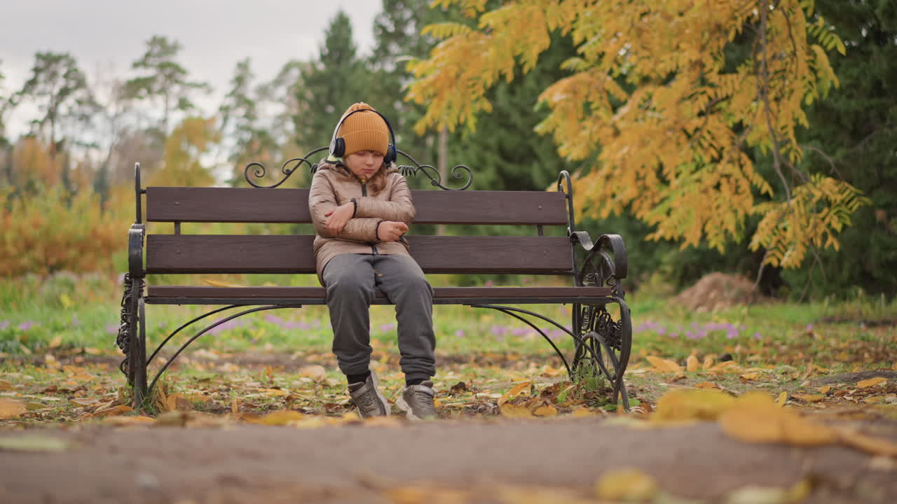 little girl seated on bench with crossed arms wearing mustard beanie and headphones listening to music amid golden autumn leaves fluttering around her as wind softly blows