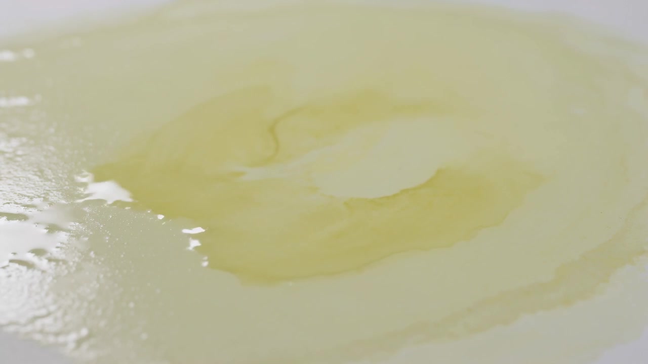Pozzanghera giallo pallido che si espande formando anelli su una superficie bianca liscia, con riflessi modellati dalla tensione.