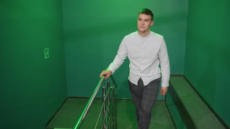 front shot of male in shirt and trousers walking up stairs, moving into billiard room. green lighting casts a unique glow on surroundings. confident expression and casual stride