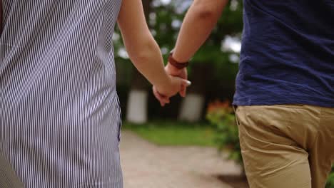Close-up-footage-of-a-young-couple-holding-hands.-Walking-on-a-green-street-together.-Bright-day.-Backside-view