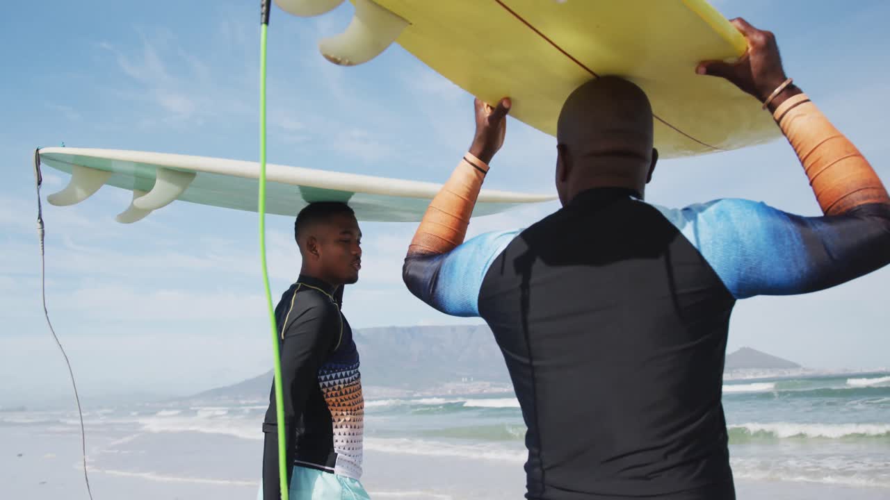 padre afroamericano y hijo adolescente de pie en la playa sosteniendo tablas de surf en la cabeza y hablando