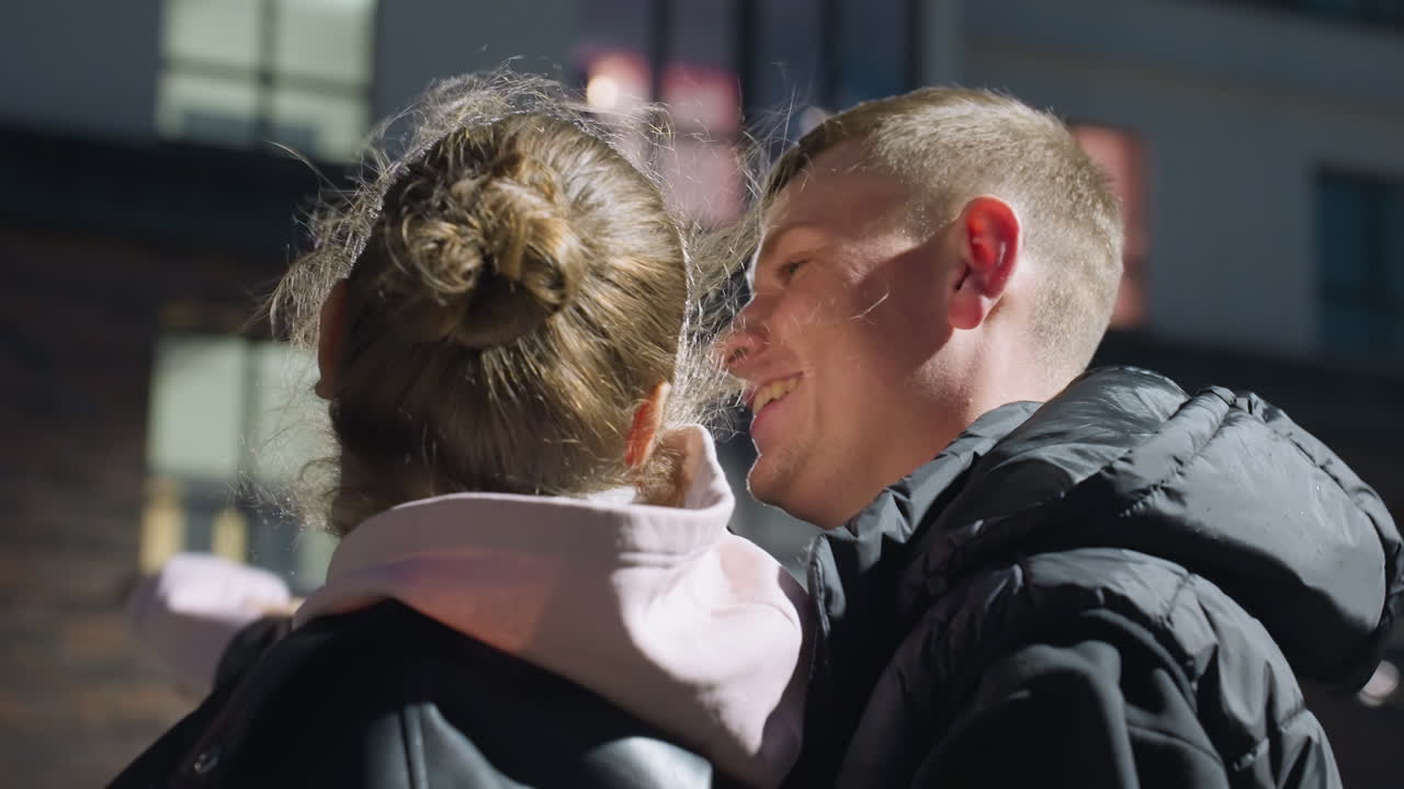 Young adults dancing closely with smiles as evening light reflects off their jackets, wind plays with hair, and modern building windows glow in background