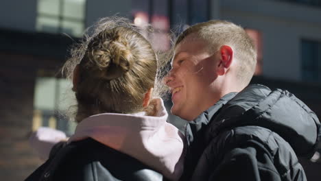 young adults dancing closely with smiles as evening light reflects off their jackets, wind plays with hair, and modern building windows glow in background