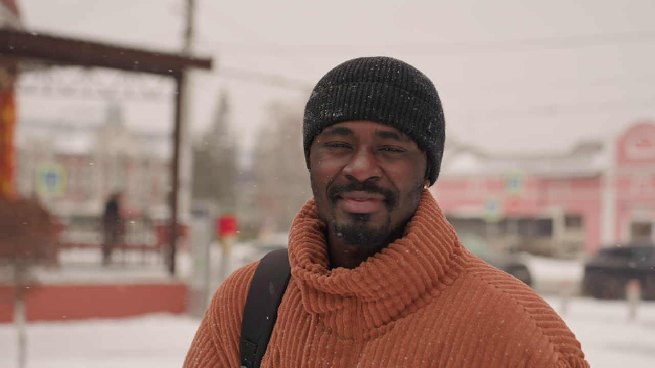 Uomo di colore che sorride nella neve vicino a un chiosco del mercato, che indossa un maglione arancione spesso e un berretto nero, tracolla dello zaino sulla spalla, passeggiata spontanea con leggera nevicata sul cappello, sorriso caloroso, edifici urbani color pastello