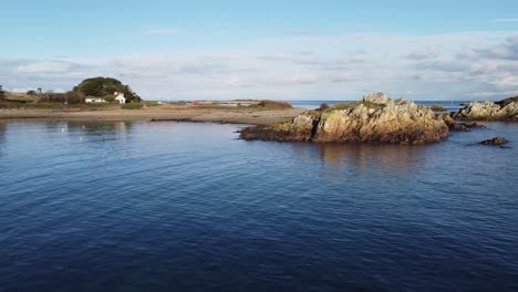 Low-drone-flight-across-Bordeaux-harbour-at-high-tide-landward-towards-beach-and-beach-side-house-on-a-calm-sunny-afternoon-in-Guernsey