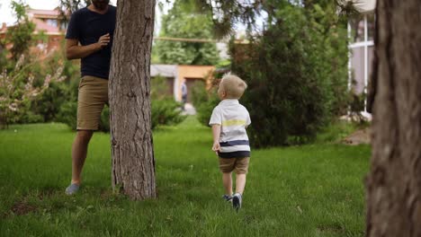 a little boy is playing with his father in hide and seek in the garden. he is happily running on the grass. the father catches his son, raises to his hands and turns him around. slow motion