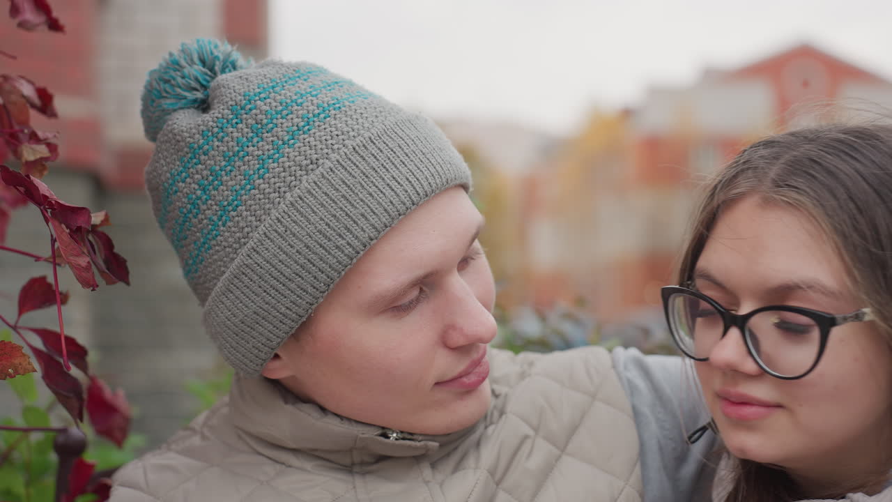 Close up of husband in knitted cap seated outdoors embracing wife closely as she rests her head on his chest, surrounded by autumn leaves and urban backdrop
