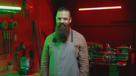 portrait shot of caucasian youngmale welder with long beard, wearing apron, crossing hands and looking at camera in workshop