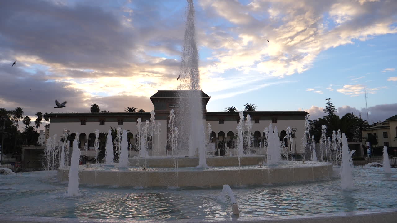 Gran fuente de agua en una plaza urbana con un edificio majestuoso y palmeras