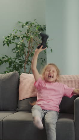 girl feels happy to beat little brother playing video game with console. emotional preschooler jumps while upset toddler frowns sitting on sofa