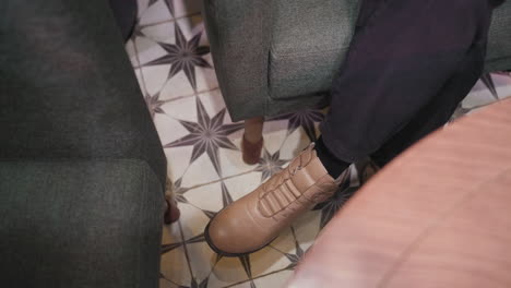 close-up view of female leg in stylish brown boot swinging slightly over patterned tile floor, seated on cushioned chair in cozy setting, soft lighting enhances modern fashion