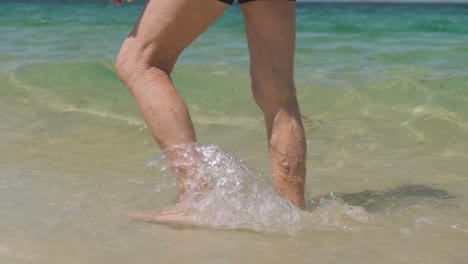 old man's legs walking on beach shore, clear ocean waves splashing