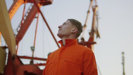 Hübscher-Junger-Containerlagerarbeiter-In-Orangefarbener-Uniform,-Der-Neben-Dem-Schiff-Im-Hafen-Steht-Und-Nach-Oben-Schaut.-Großer-Kran-Am