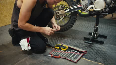 Close-up shot: A man in a gray T-shirt repairs and inspects his motorcycle, which he lifted with a jack in a garage workshop. A mechanic selects the tool he needs from a whole set of tools