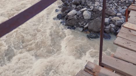 Walking-on-old-wooden-bridge-above-wild-glacial-river-in-Caucasus-mountains,-point-of-view