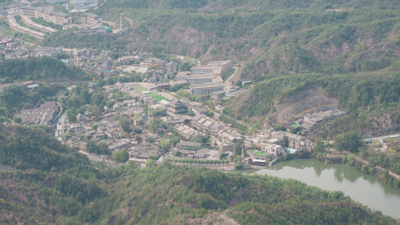 Bacino di un villaggio visto dall'alto con stagno fluviale, denso gruppo di case accanto a un corso d'acqua serpeggiante, strade e terrazzamenti collinari, con agricoltura su piccola scala e mercati locali impliciti, prospettiva di mappatura da drone, comunità.