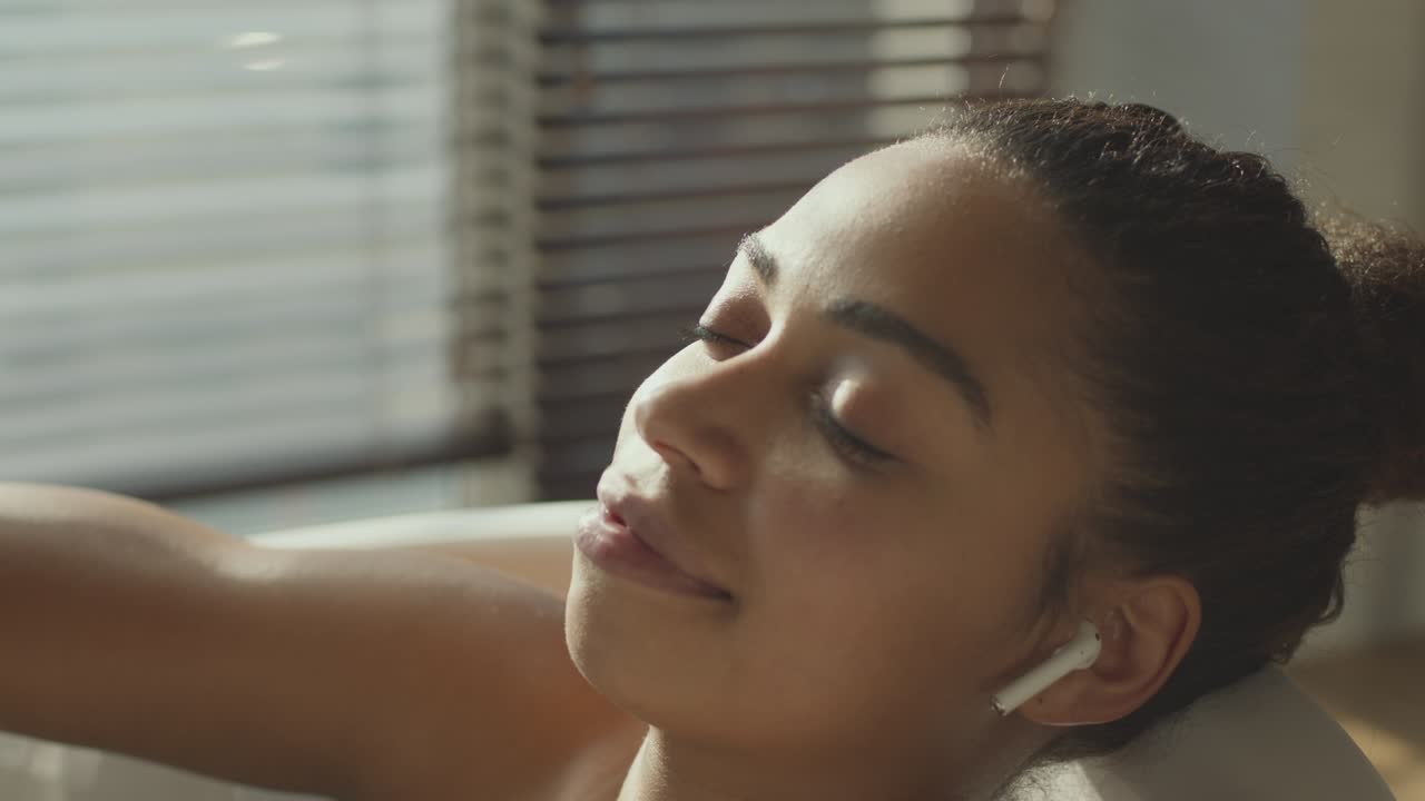 близкий портрет спокойной расслабленной афроамериканской женщины, наслаждающейся музыкой через наушники, отдыхающей в ванне с закрытыми глазами