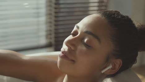 close up portrait of calm relaxed african american woman enjoying music via earphones, resting in bath with closed eyes