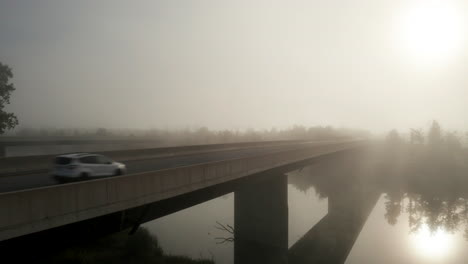 ascending aerial view of cars on highway bridge crossing calm river as morning light shines through dense fog hanging over the landscape