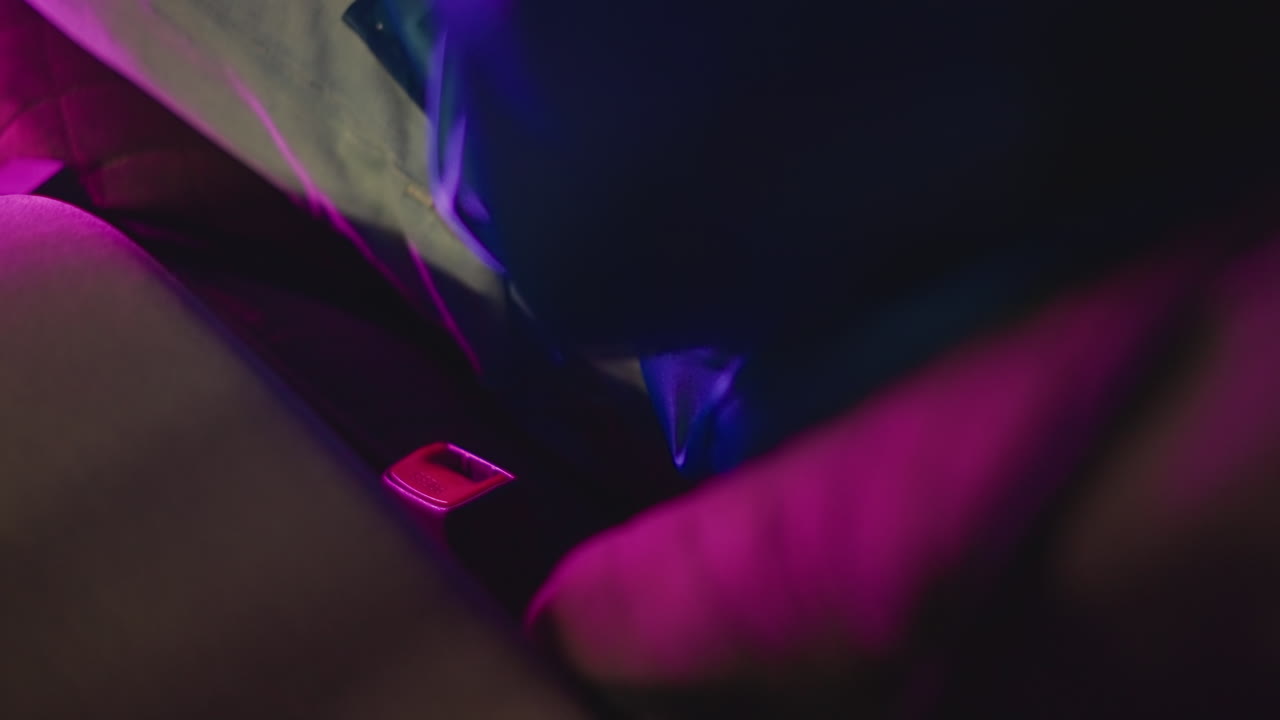 Pasajero en el asiento trasero de neón abrochándose el cinturón, mano ansiosa buscando la hebilla bajo luces magenta y añil, primer plano sincero del pestillo y la correa dentro de un taxi urbano, momento de tensión con poca luz para un viajero