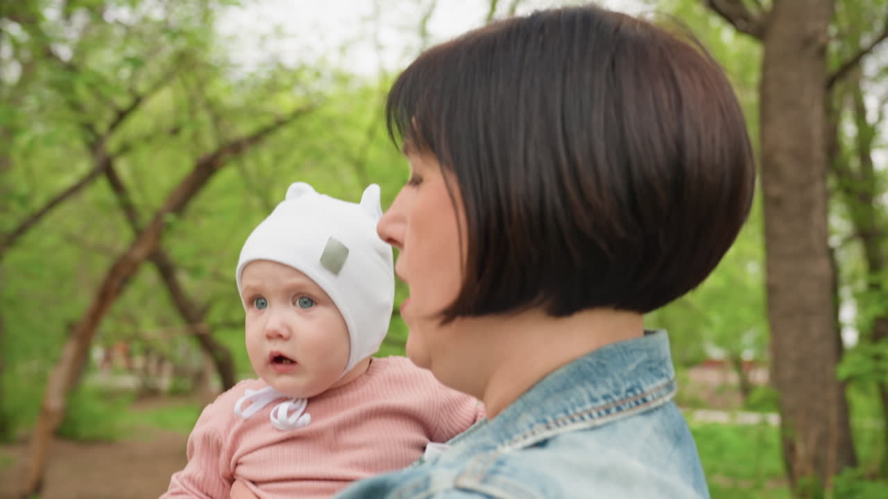 Gentle Woman Walks With Baby Amidst Trees, Serene Scene Of Mother Caring For Infant On Park Trail, Calm Moment As Woman In Denim Jacket And White Hat Strolls With Baby Through Lush Park
