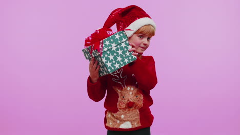divertida niña niño niño lleva suéter de año nuevo recibió un regalo, interesado en lo que dentro de la caja de regalos