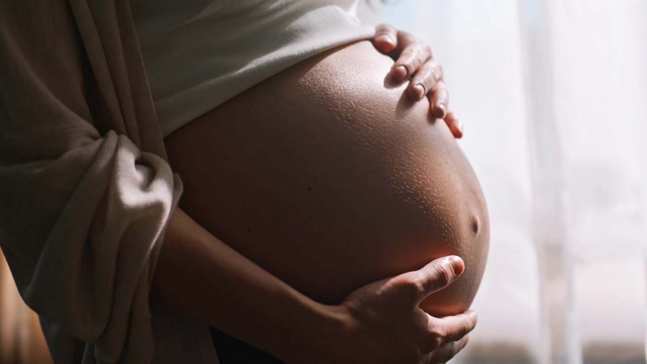 Madre in attesa che accarezza il pancione vicino a una finestra luminosa. Primo piano delle mani sul pancione.