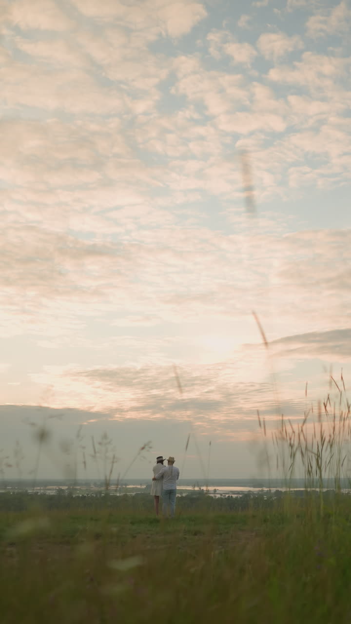 Partners stand on a grassy hill beside a lake at sunset, gazing at the sky. The man, in a white shirt, hat, and jeans, lovingly holds the woman by the waist. She wears a black hat and a white dress
